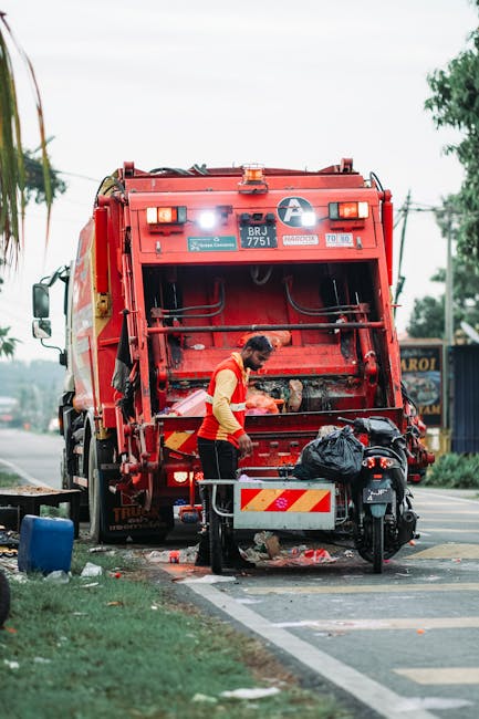 A small, old blue pickup truck is parked on the side of a street, loaded with a large quantity of mixed waste materials including cardboard boxes, plastic bags, and various scrap items. The truck’s 