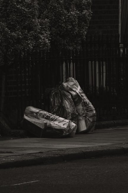 Three large black plastic rubbish bags filled with discarded waste are placed on the pavement beside a metal fence with vertical bars. The bags appear slightly crumpled and are stacked irregularly, wi