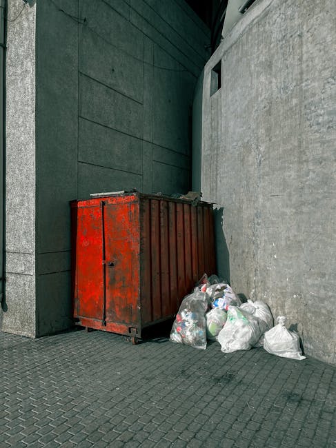Three large black plastic rubbish bags filled with discarded waste are placed on the pavement beside a metal fence with vertical bars. The bags appear slightly crumpled and are stacked irregularly, wi