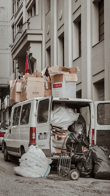 A large collection of mixed waste, including cardboard boxes, plastic bags, paper, and discarded packaging, piled outside a commercial building in an urban area. The waste is scattered on the paved gr